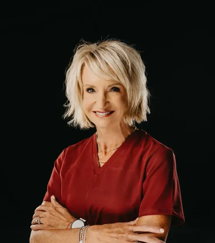 A smiling woman with blonde hair, wearing a maroon scrub top, poses with arms crossed against a plain black background.