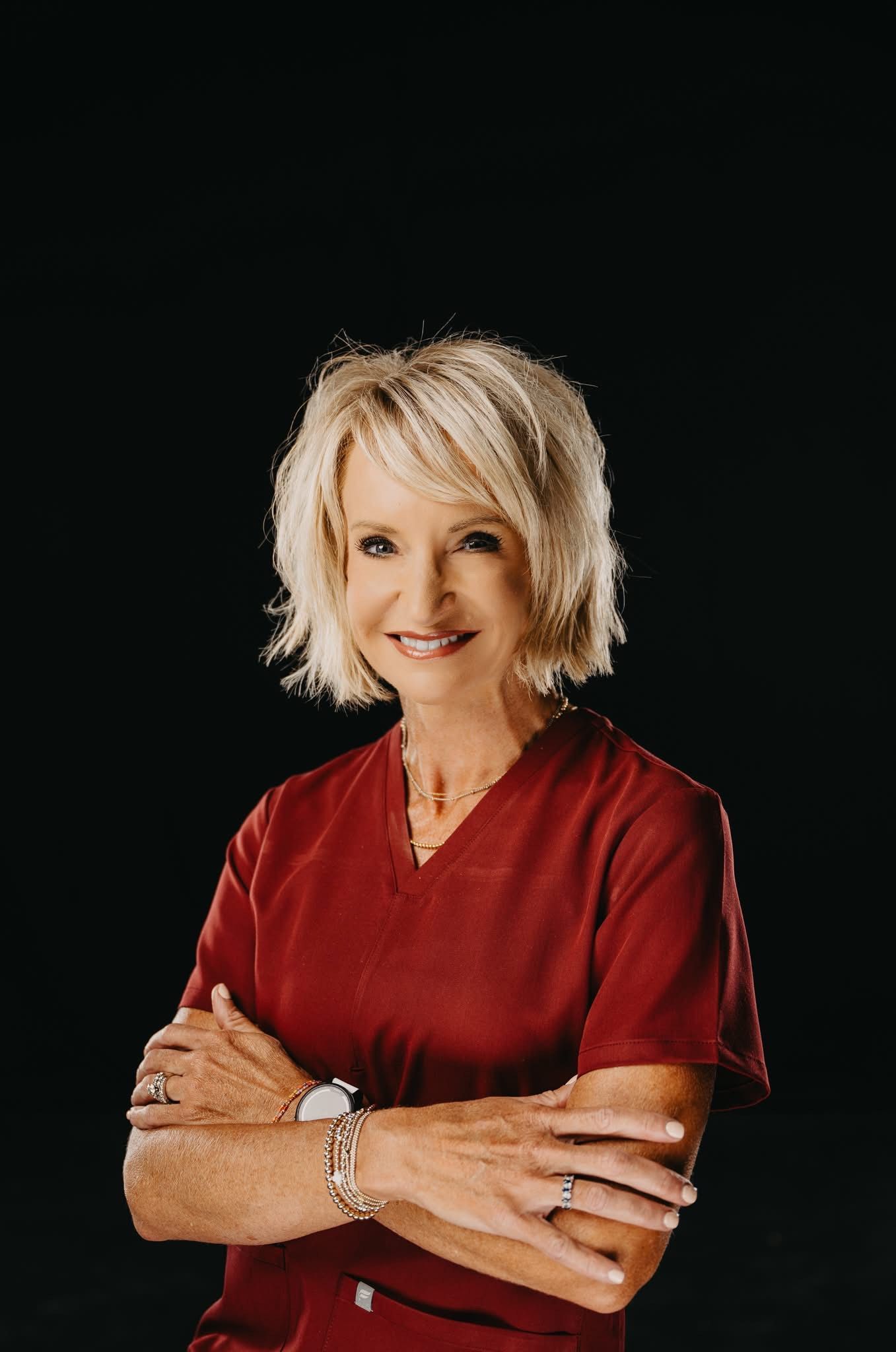 A smiling woman with blonde hair, wearing a maroon scrub top, poses with arms crossed against a plain black background.
