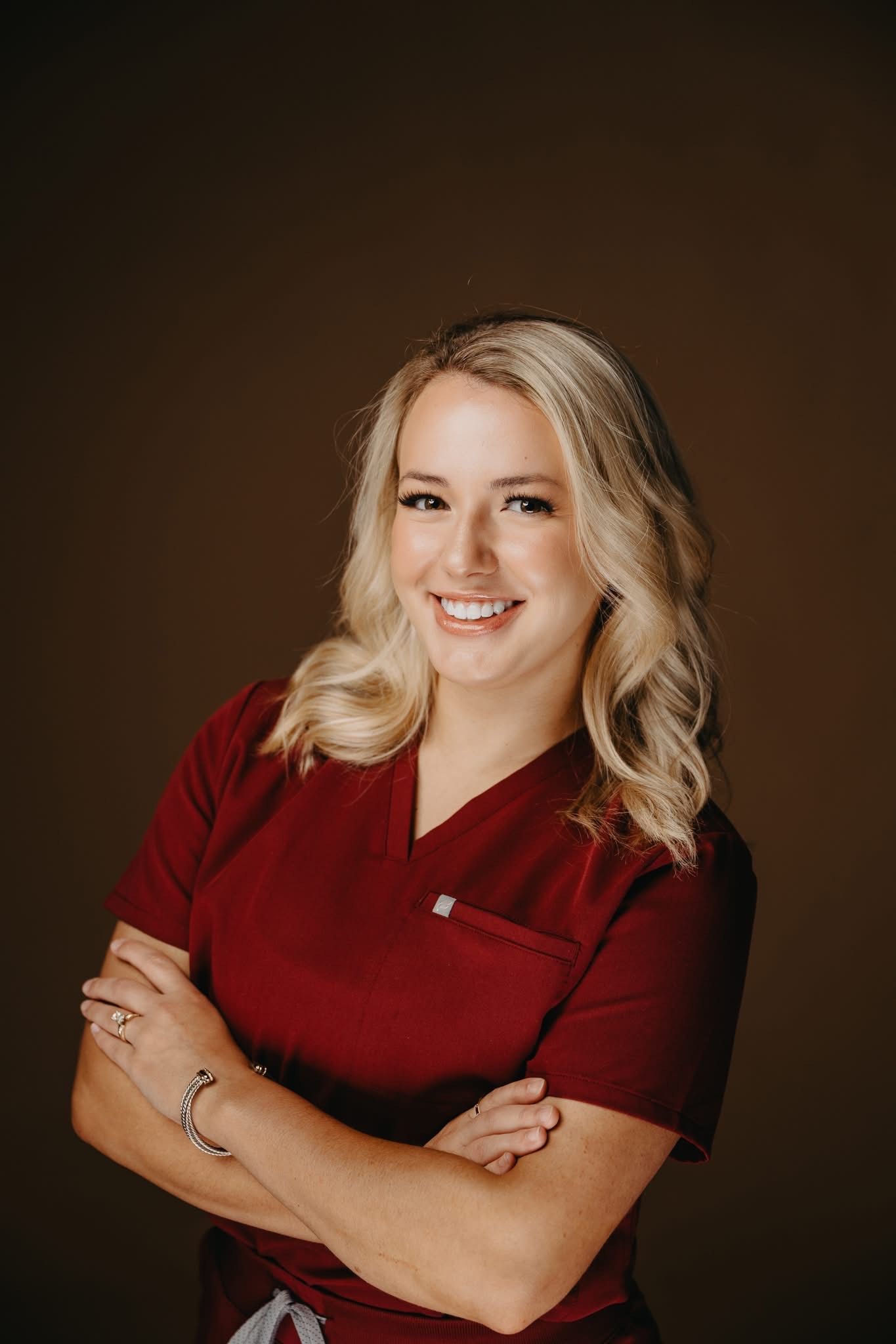 A smiling woman in a maroon medical scrub top poses with her arms crossed against a plain brown background.