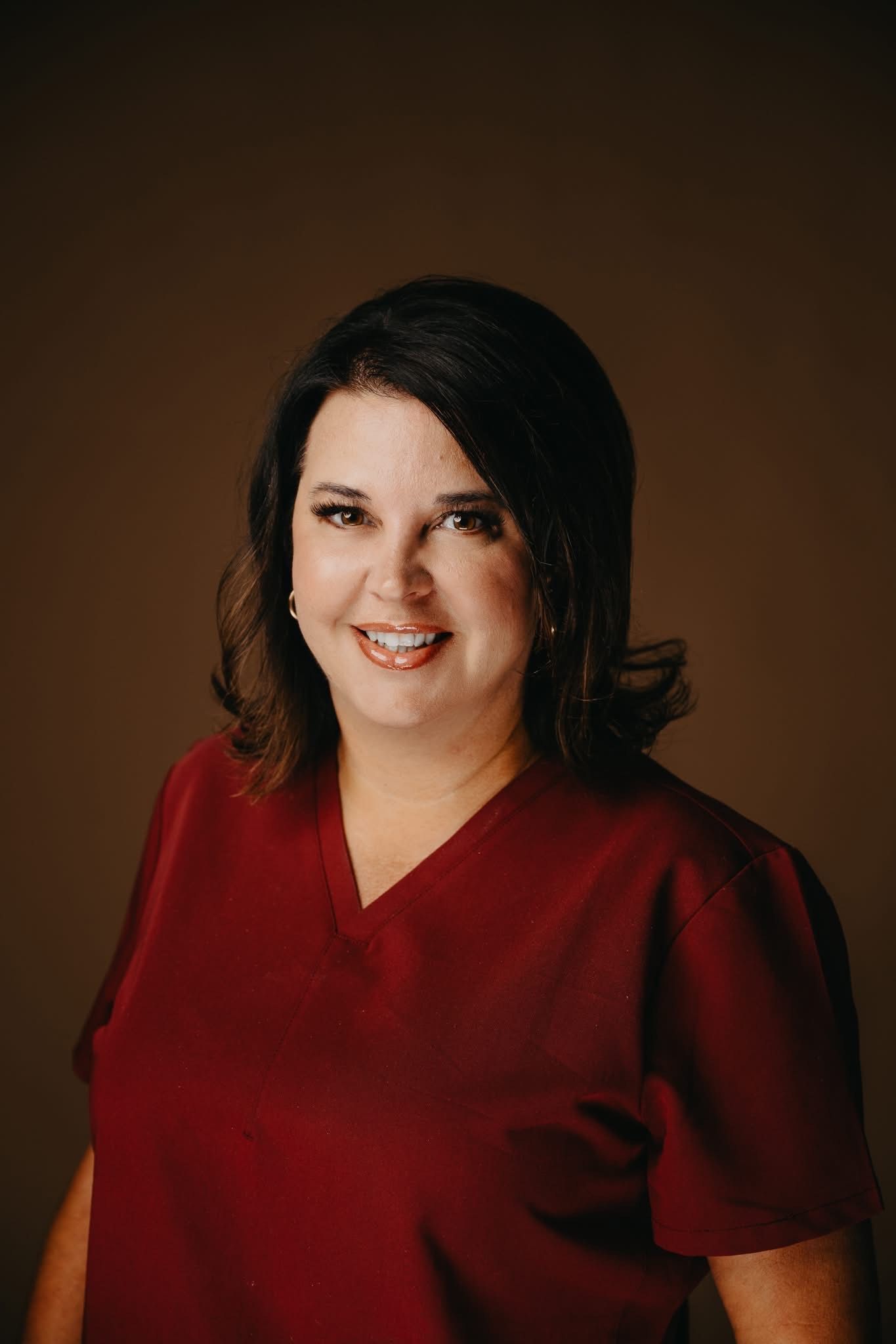 A professional portrait of a smiling person wearing a burgundy v-neck scrub top against a solid brown background.