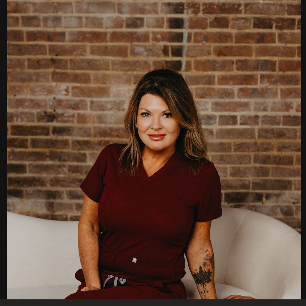 A person in burgundy scrubs with a tattoo on their arm, posing against a brick wall on a curved white sofa.