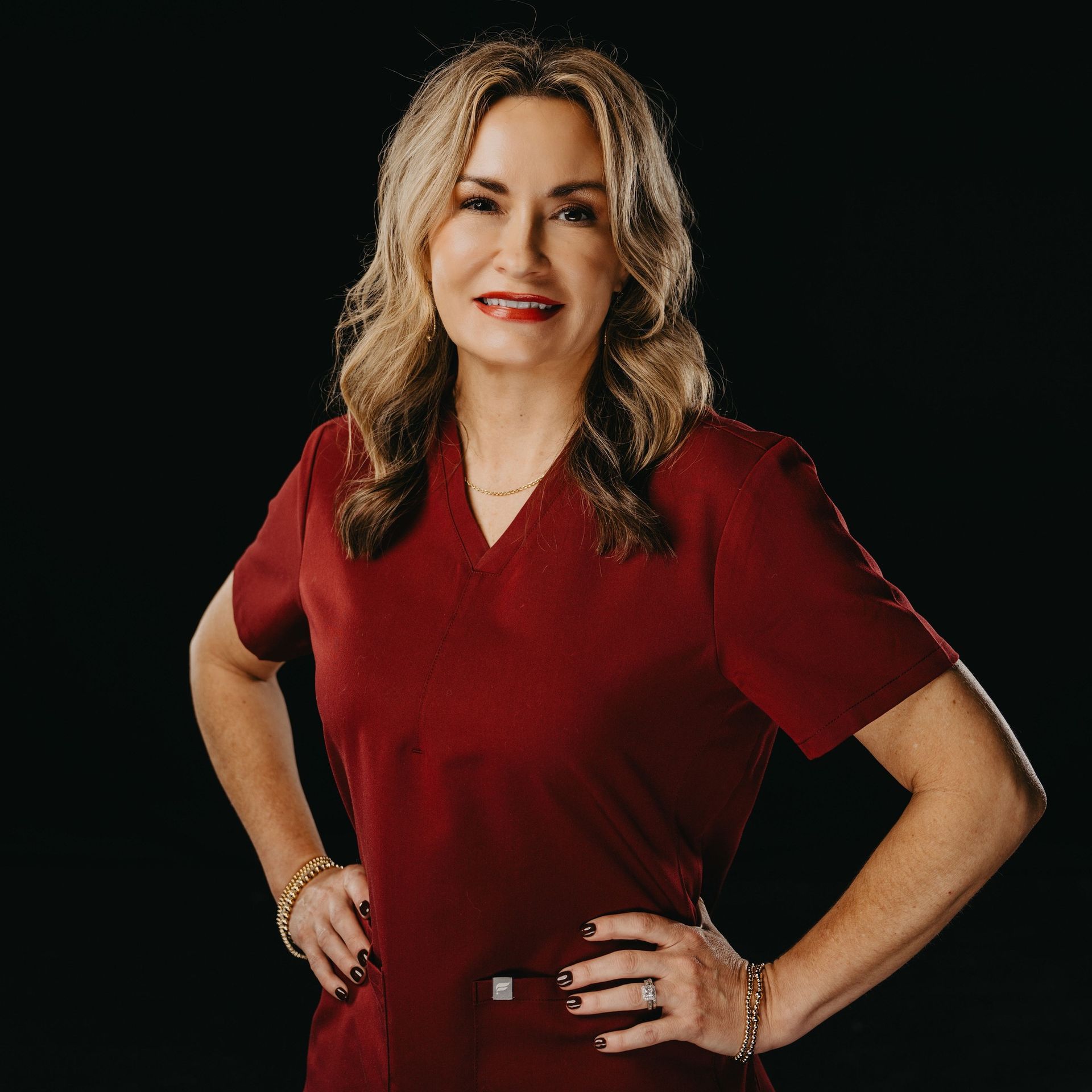 Professional headshot of a person in maroon medical scrubs, posing with hands on hips against a black background.