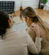 A person in a beige blazer comforts someone with long hair, placing a hand on their shoulder in a bright, modern room.