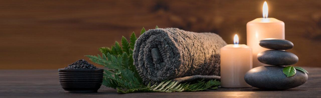 A rolled grey towel, green fern, lit candles, and stacked stones arranged on a wooden surface for a spa-like atmosphere.