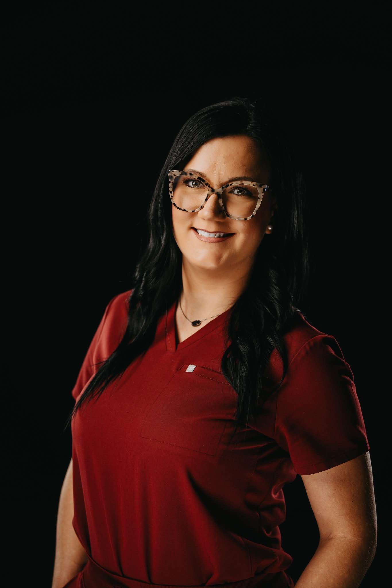 A person with dark hair and glasses wearing a maroon scrub top, smiling against a solid black background.