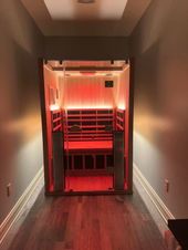 An infrared sauna glowing with red light sits at the end of a home hallway with wood flooring and light grey walls.