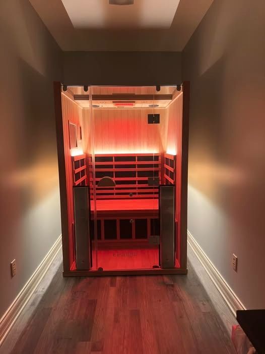 An indoor sauna with glowing red interior lights sits at the end of a narrow hallway with hardwood floors.
