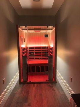 An indoor sauna with glowing red interior lights sits at the end of a narrow hallway with hardwood floors.