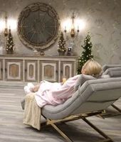 A person in a light pink robe reclines in a gray lounge chair in a decorated room with a buffet and holiday trees.