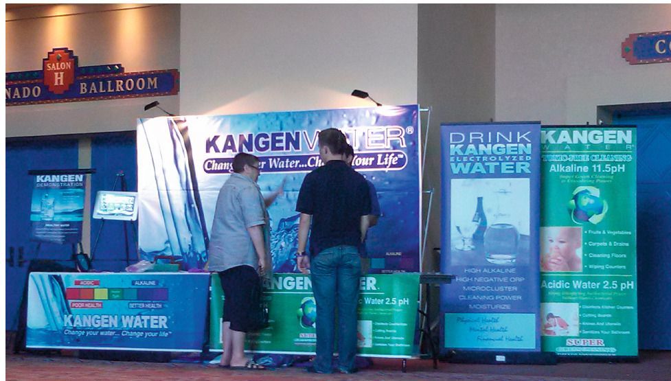 Two people at a Kangen Water display in a convention hall, with colorful banners and signs promoting alkaline water.