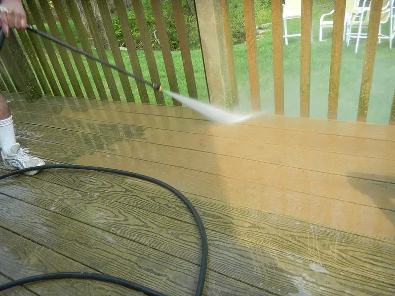Person power washing a wooden deck with a high-pressure hose, revealing a clean strip.