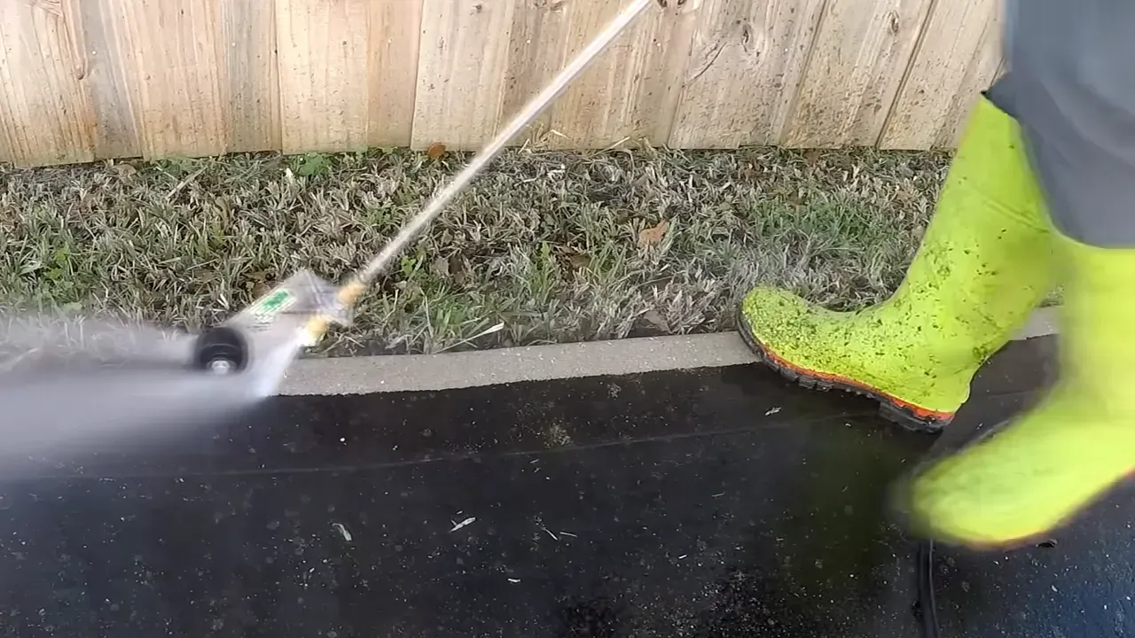 A person wearing bright green boots pressure washes a dark surface next to a fence and grass.