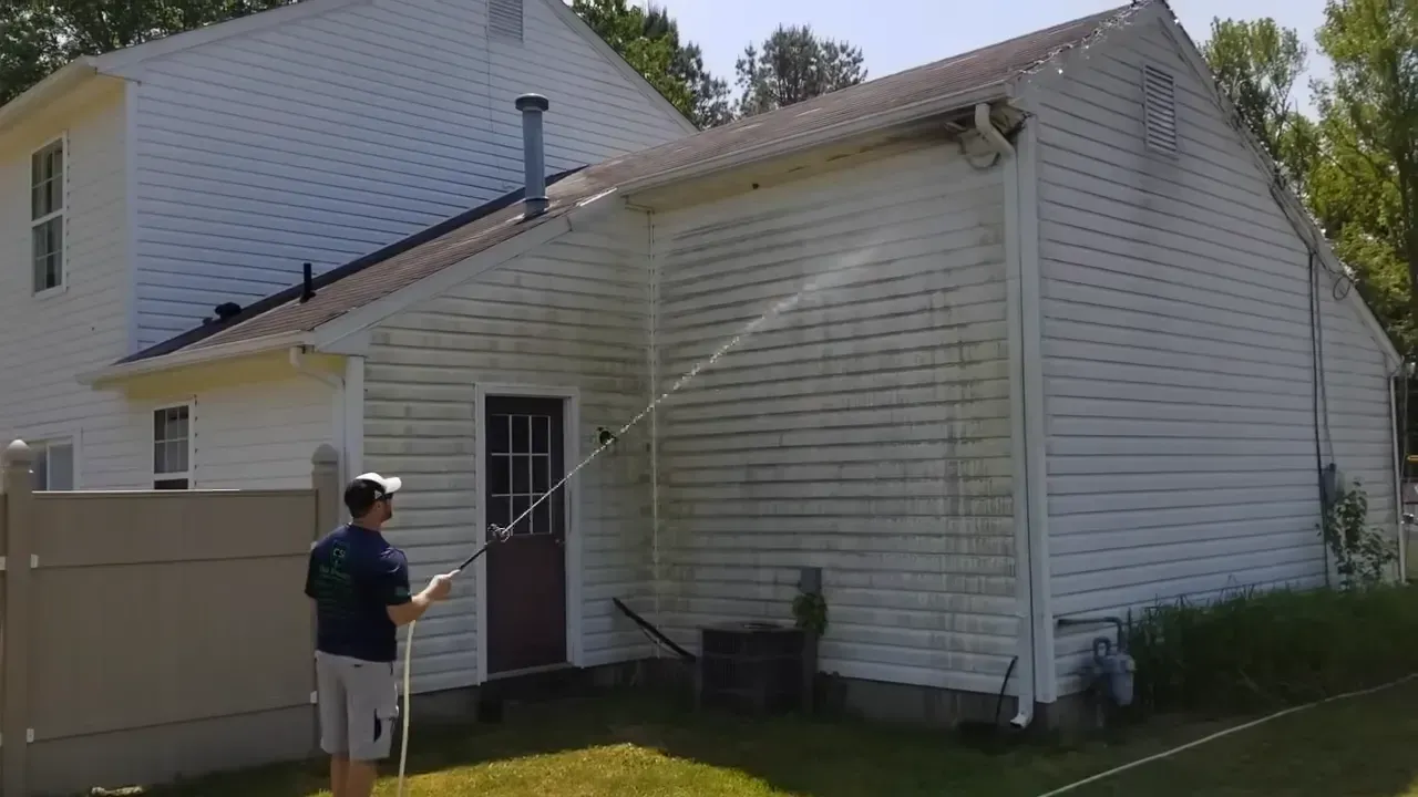 Man power washing the side of a white house with a brown door. Green grass and blue sky.