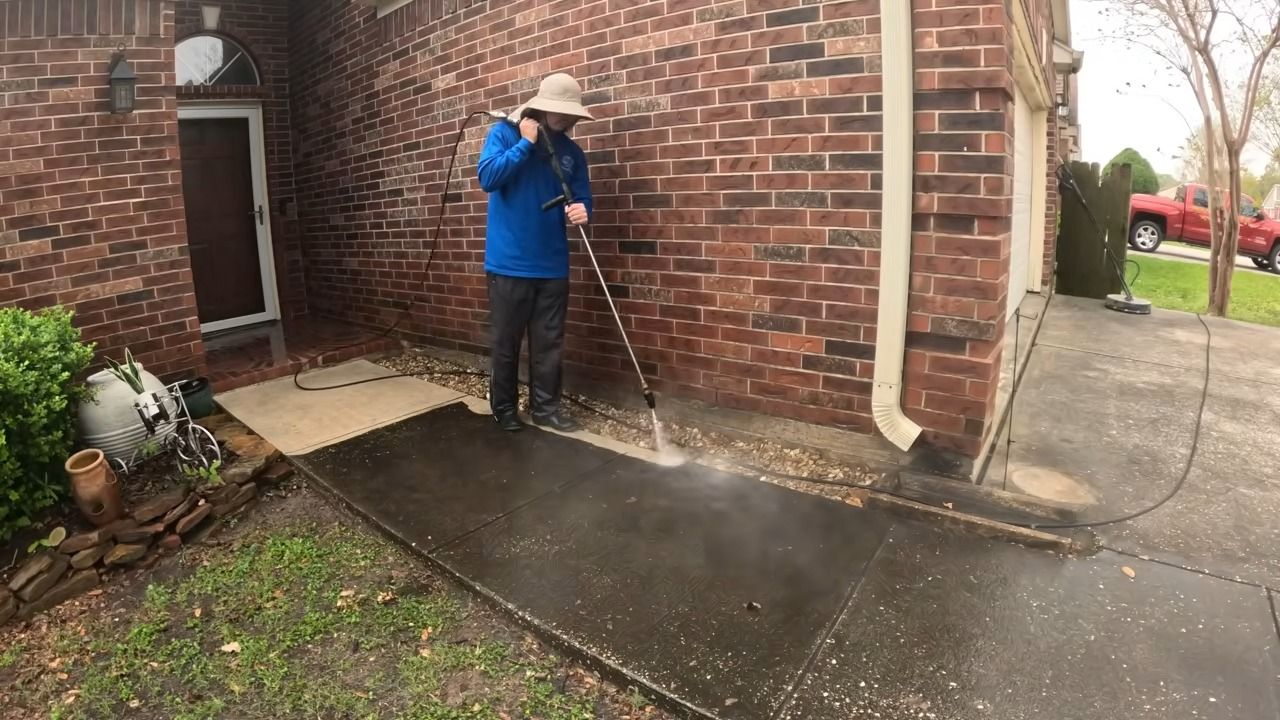 Person pressure washing a dark concrete walkway next to a red brick building.