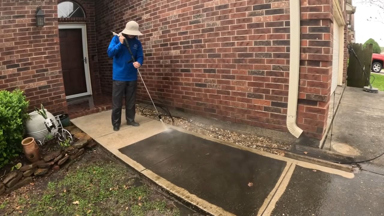 Person power washing a concrete walkway in front of a brick house.