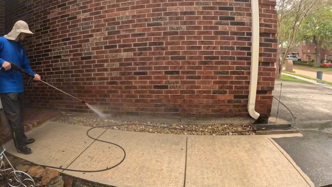 Person power washing a brick building's exterior. The walkway and ground around the foundation are being cleaned.