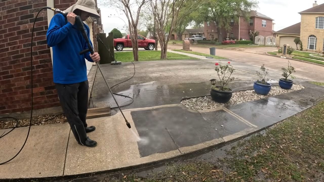 Man in blue jacket power washes a concrete driveway next to rose bushes in front of a house.
