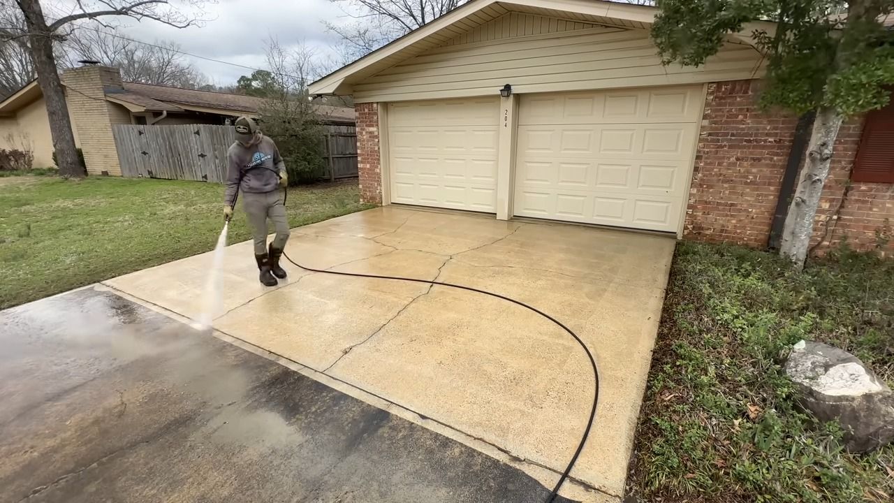 Person power washing a driveway in front of a tan garage. The driveway is wet, and the sky is overcast.