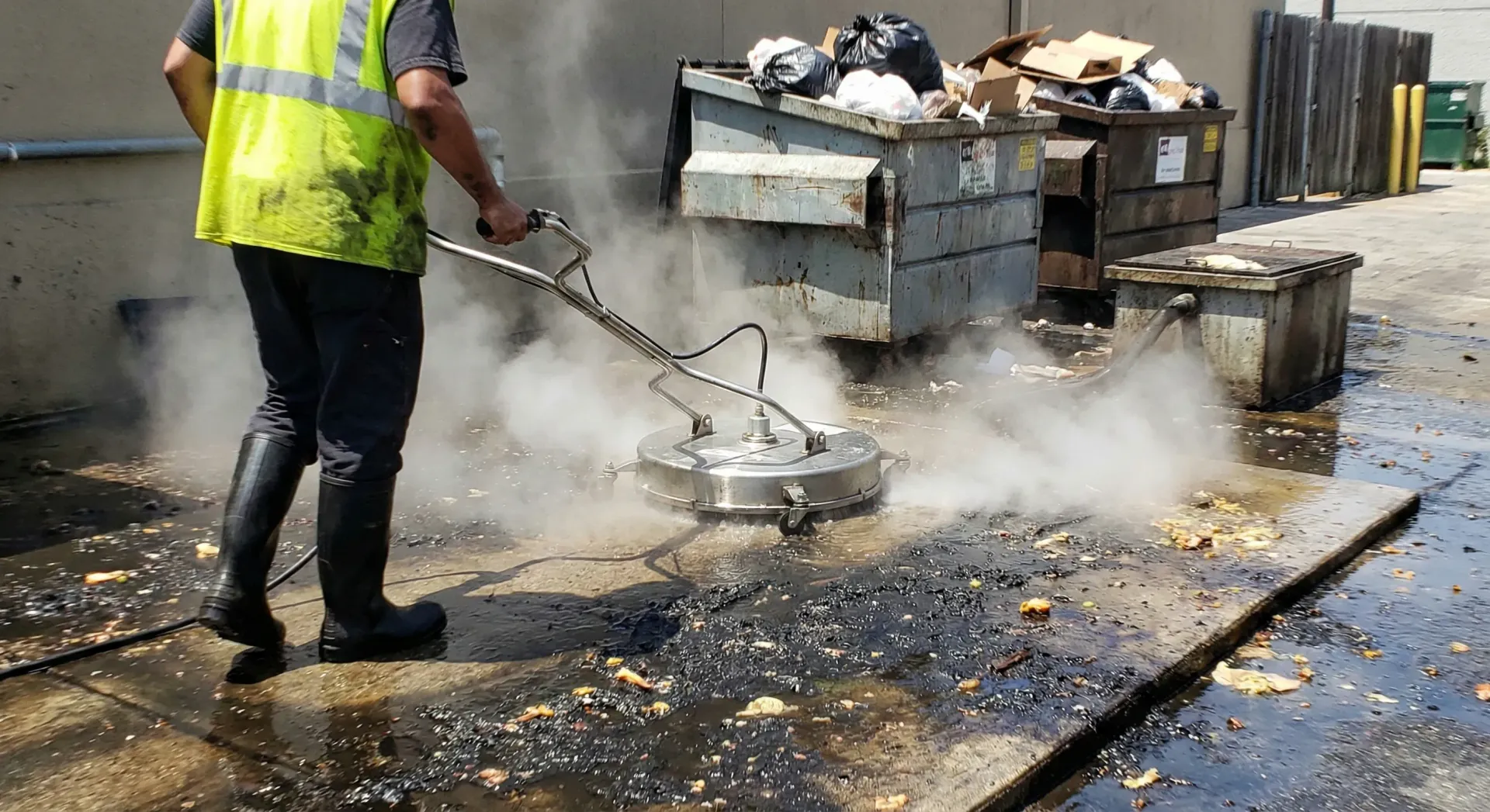 Man power washing a concrete surface near dumpsters, creating steam.