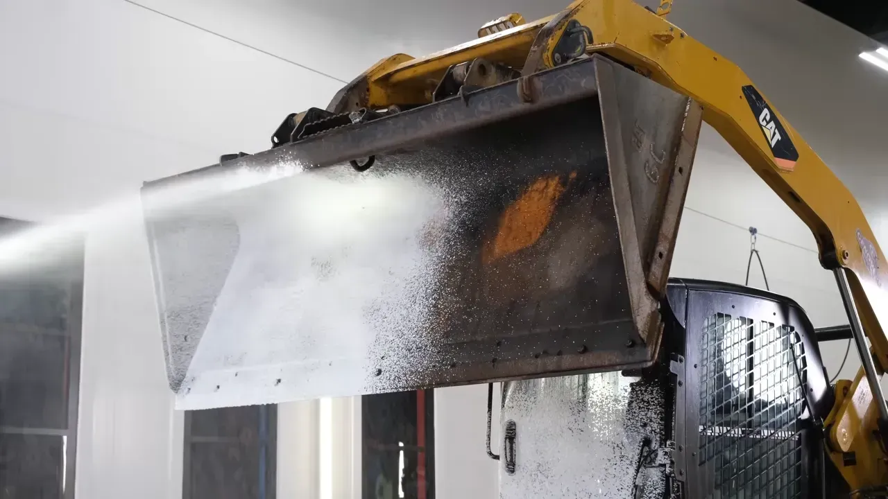 A yellow skid steer washing the bucket with water, inside a building.
