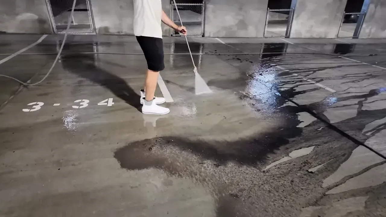 Person spraying water on a concrete floor in a parking garage. The ground is wet.