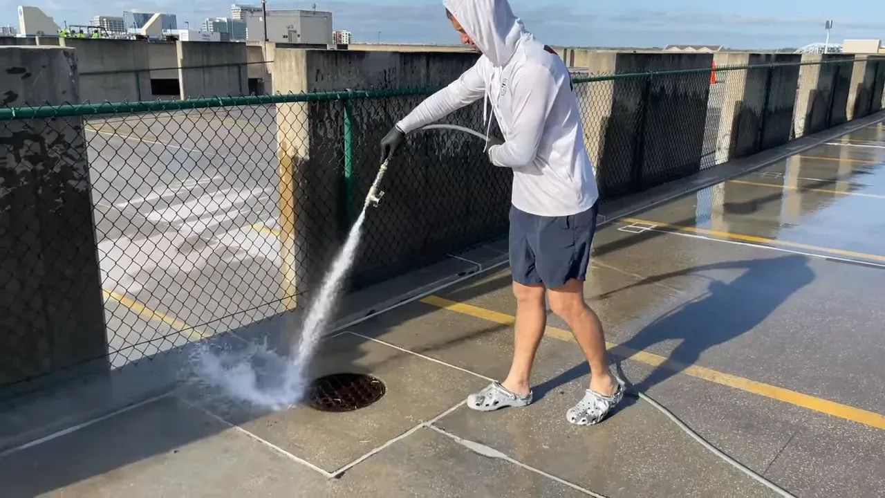 Person wearing a hoodie and shorts washing a parking garage floor with a hose.