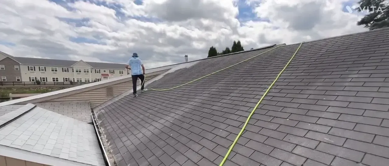 Person walks on a shingle roof with a safety rope, houses in the background on a cloudy day.