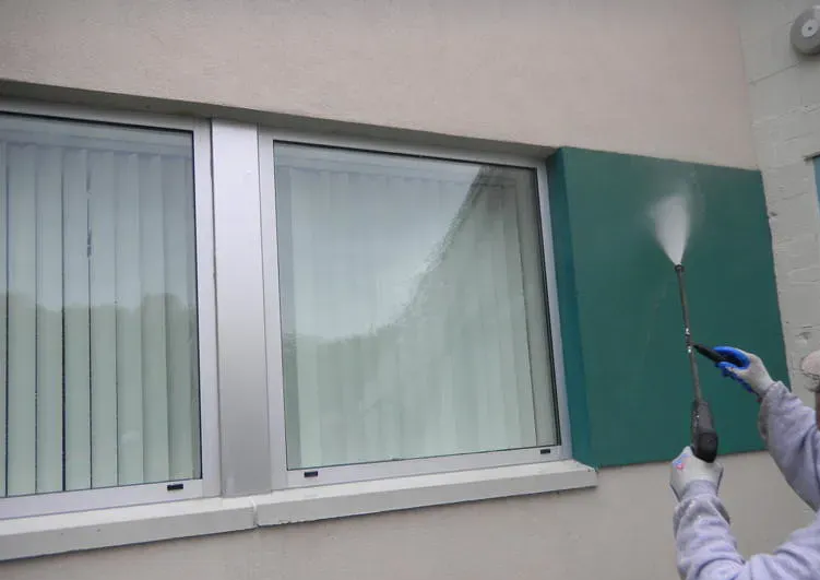 Person power washing a teal wall next to a window with white blinds.