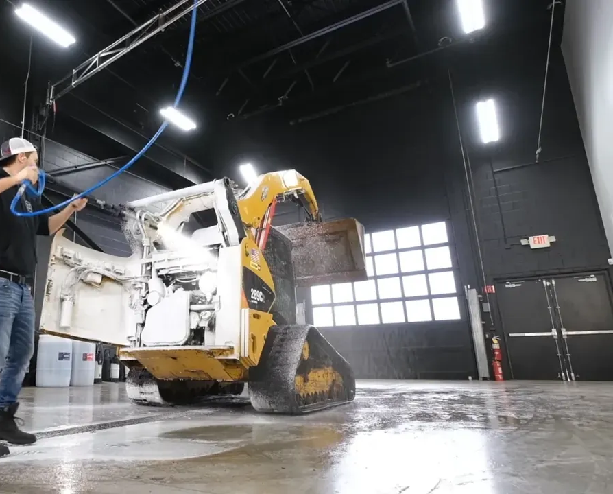 Man washing a yellow skid steer loader with a hose in a large garage.