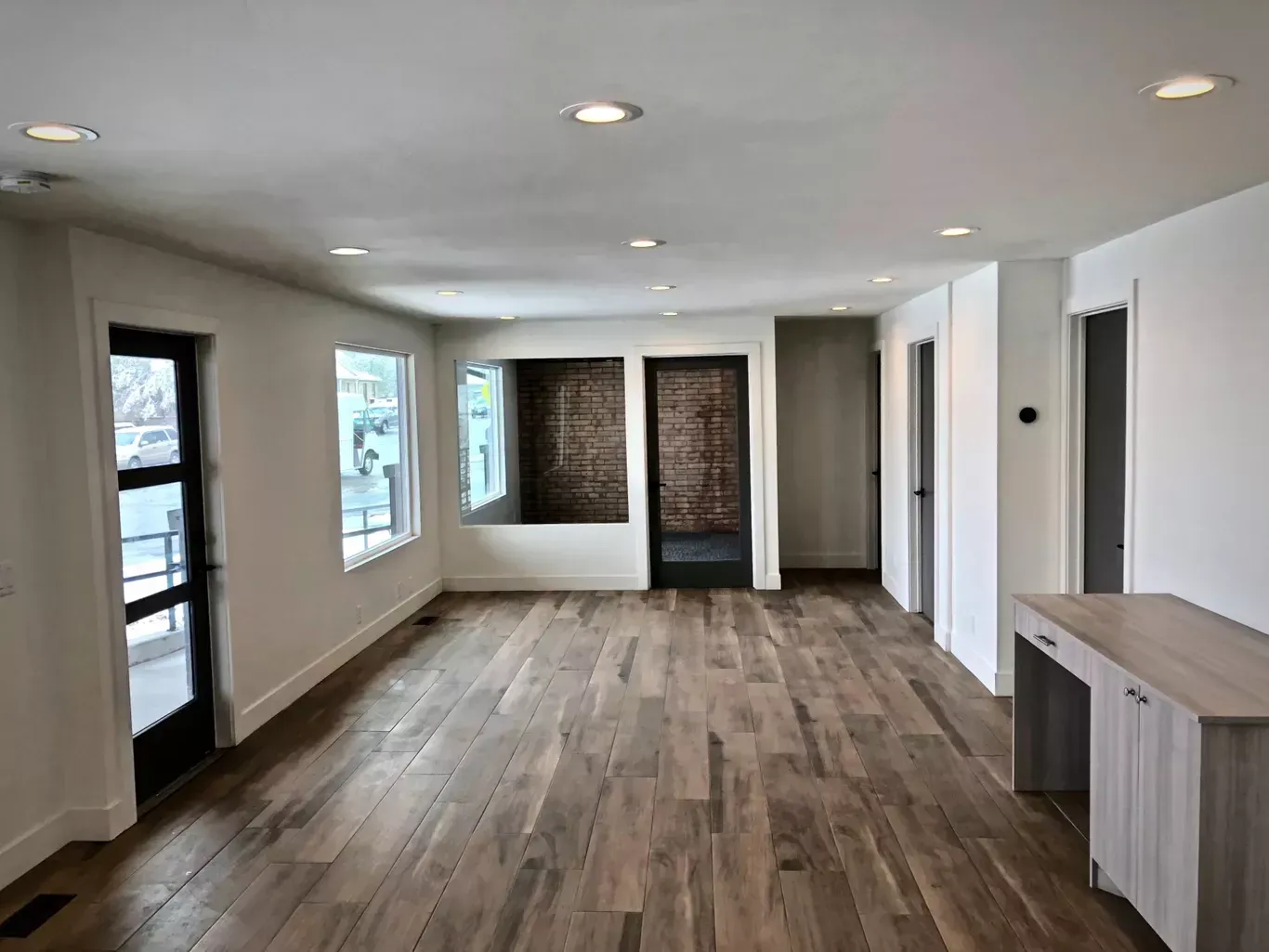 Empty room with wood floors, white walls, and recessed lighting. A dark door and brick wall accents.