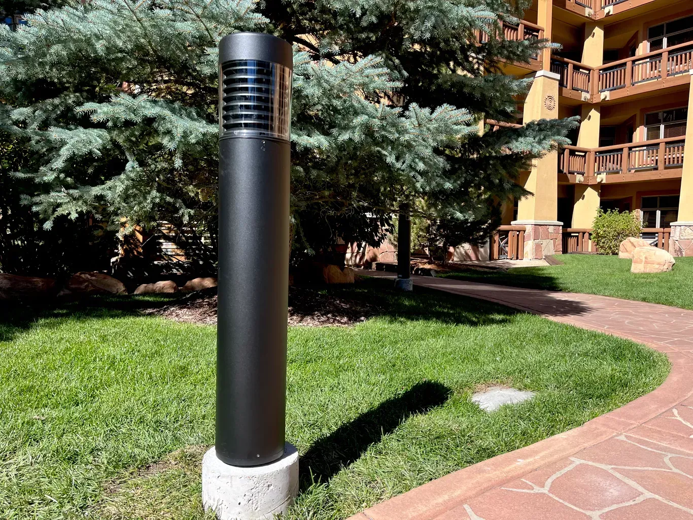 Black cylindrical bollard light on concrete base beside a red brick walkway and green grass.