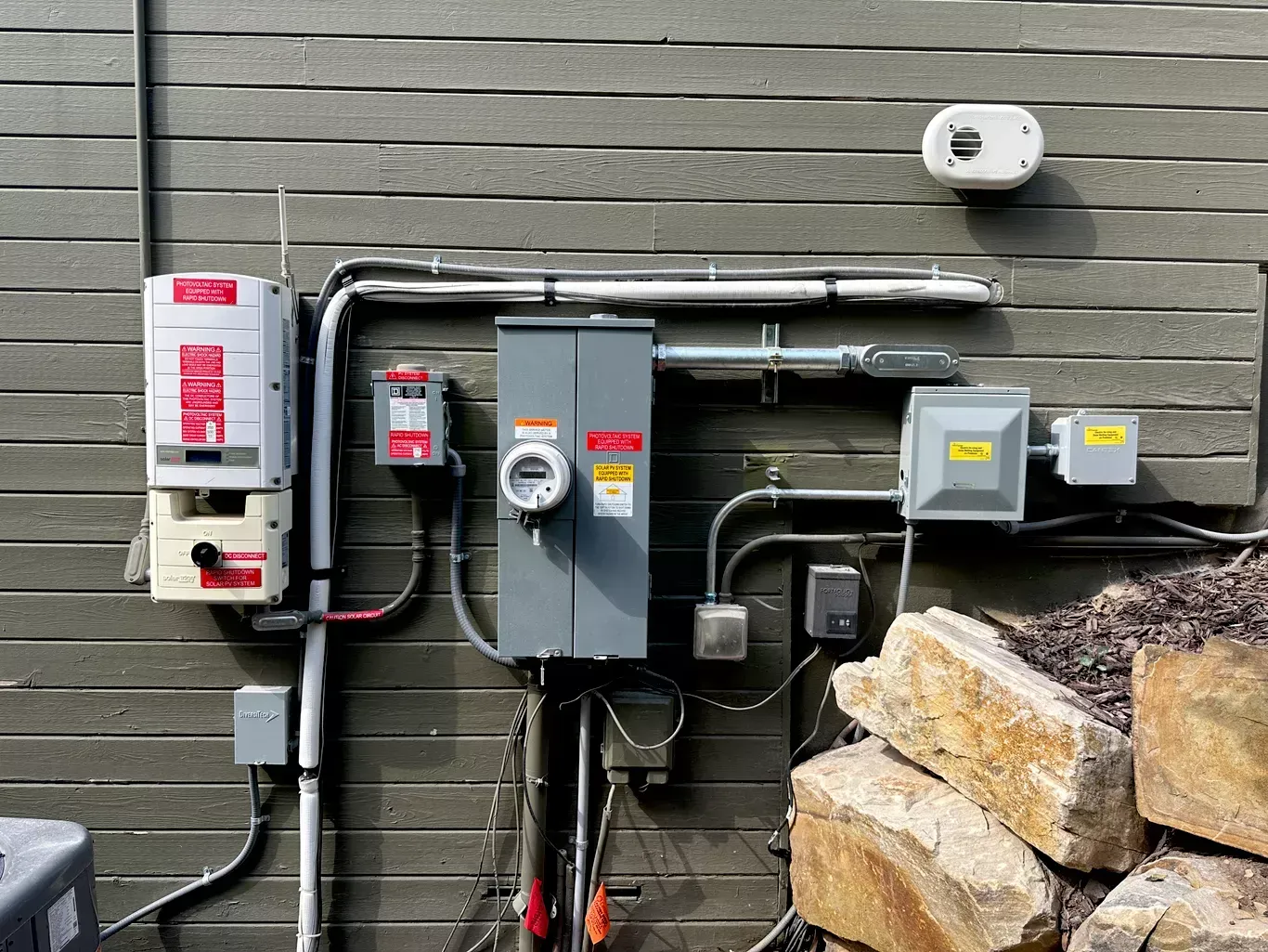 Solar panel electrical components mounted on a grey siding wall. Includes a power inverter, breaker box, conduit, and surge protector.