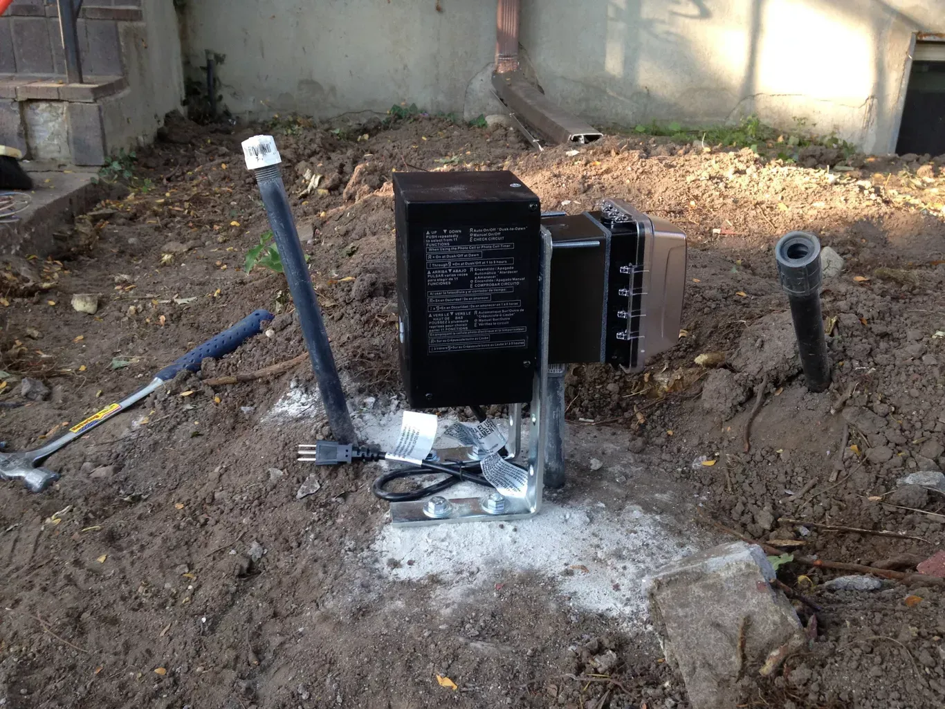 Black transformer box on a concrete base in a dirt yard with tools and pipes.