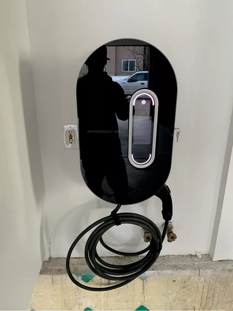 Black electric vehicle charger mounted on a white wall with its cable coiled below. A shadowy reflection of a person is visible within the charger's reflective surface.
