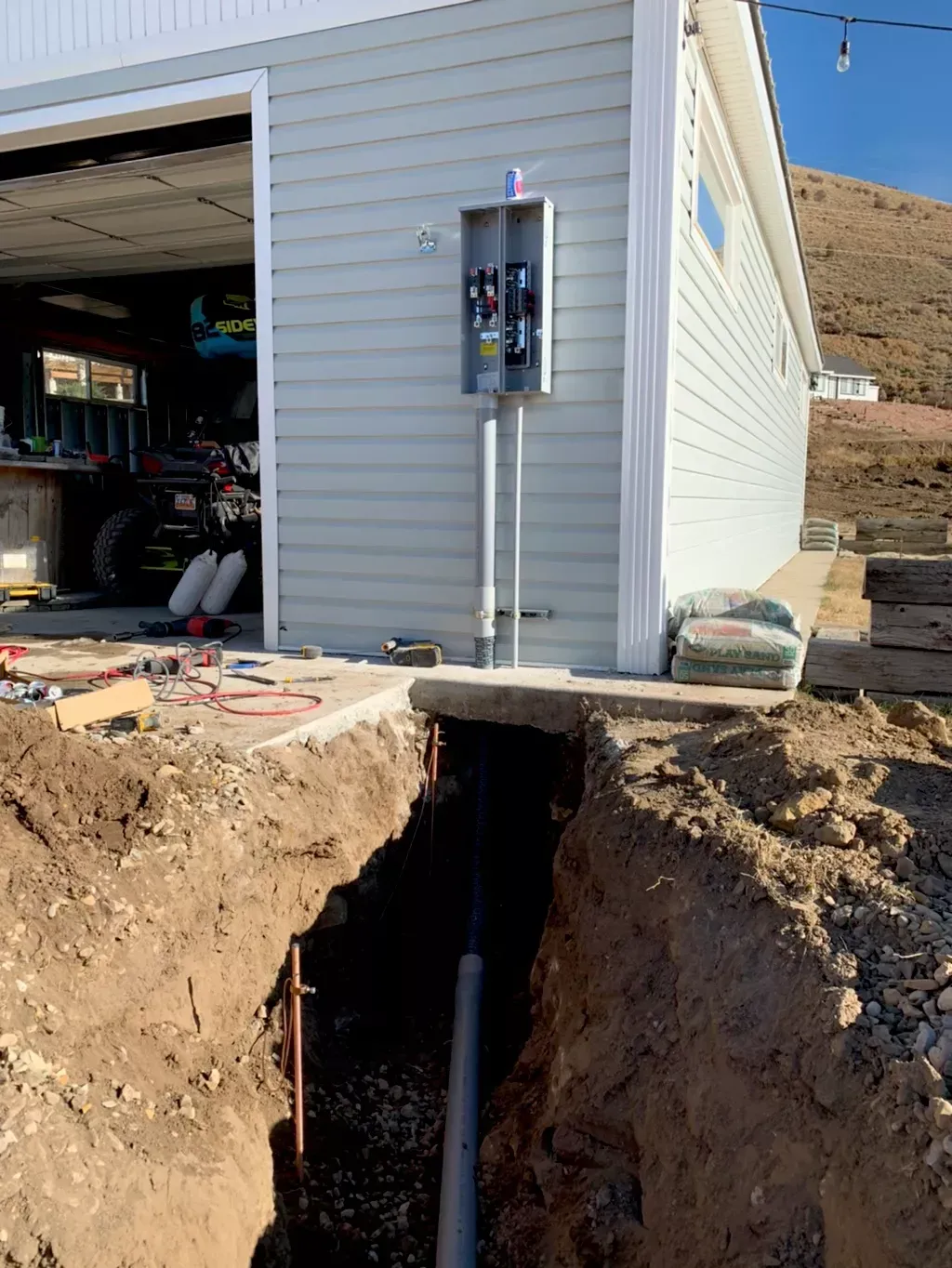 A trench dug for electrical work with a metal electrical panel mounted on the side of a light blue building.