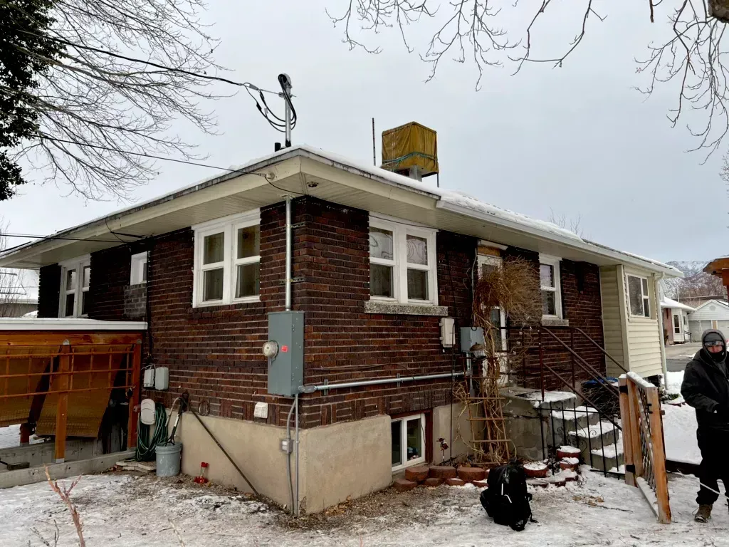 Back of a brick house covered in snow; water tank on roof, person standing to the right.