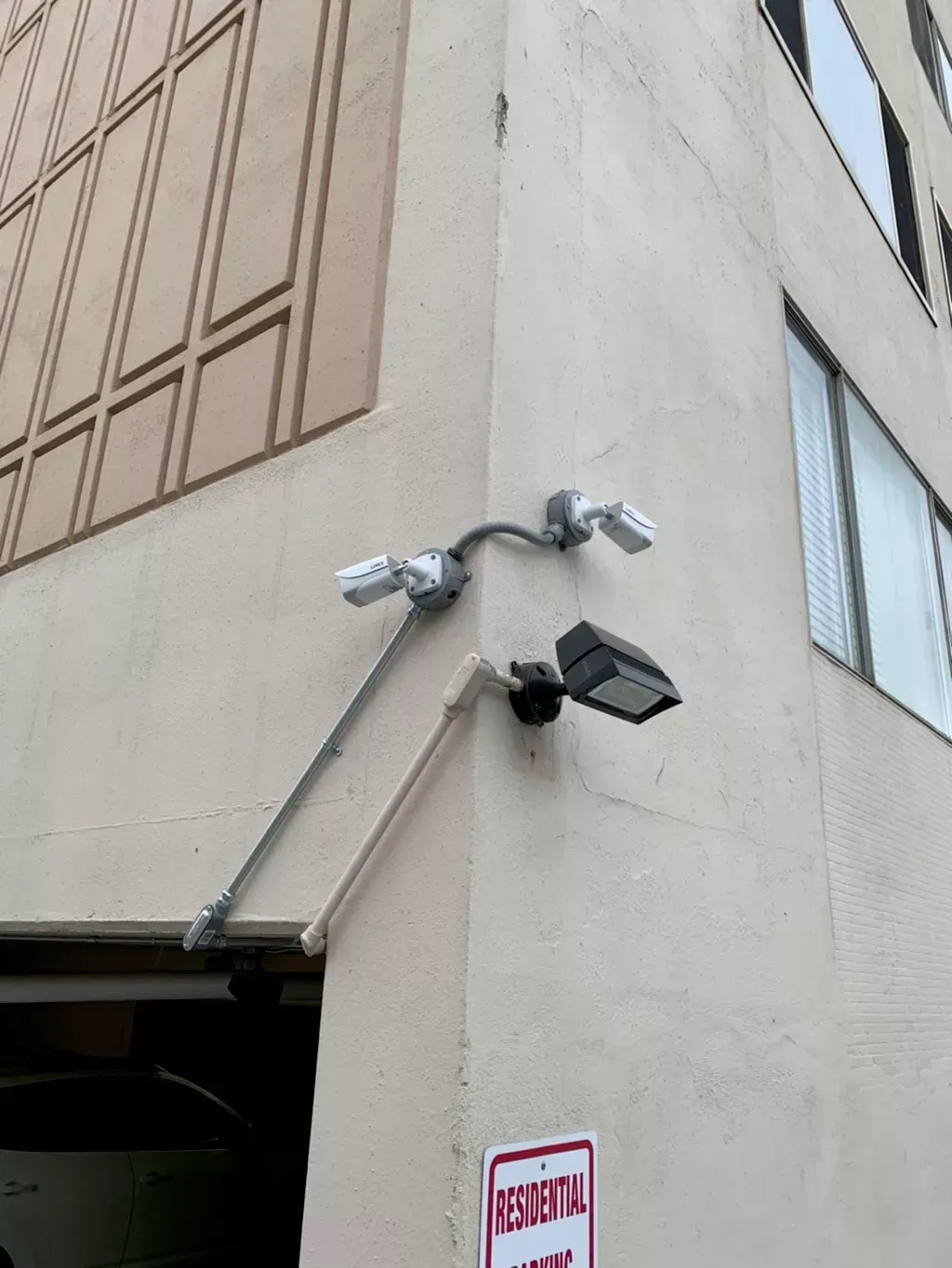 Corner of a building with security cameras and a parking sign; the building is tan with windows and a garage entrance.