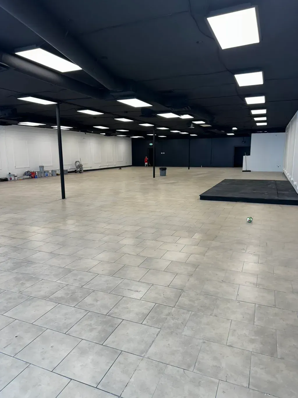 Empty, open indoor space with light-colored tile floor, dark ceiling, white walls. A small black stage is in the foreground.