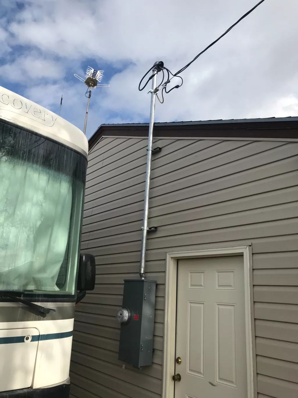 RV parked next to a building with electrical meter and service drop. Cloudy sky in background.