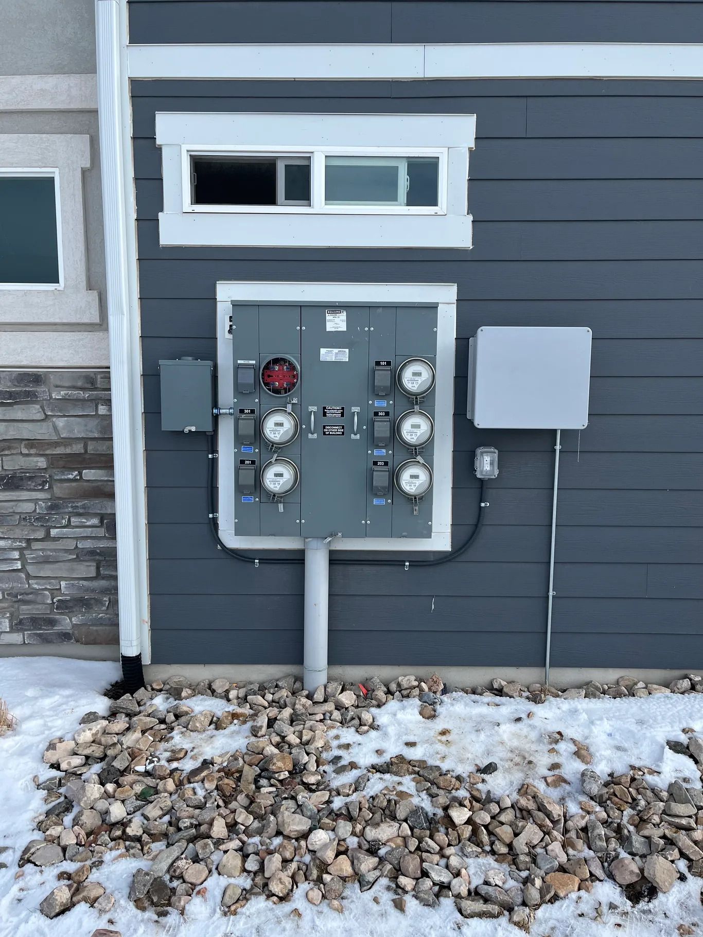 Gray electrical panel on a gray house exterior with multiple meters and conduit, set in a snowy, rocky yard.