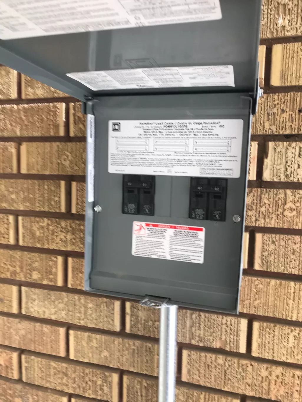 Exterior electrical panel with two circuit breakers mounted on a brick wall. The panel door is open, and a metal pipe supports it.