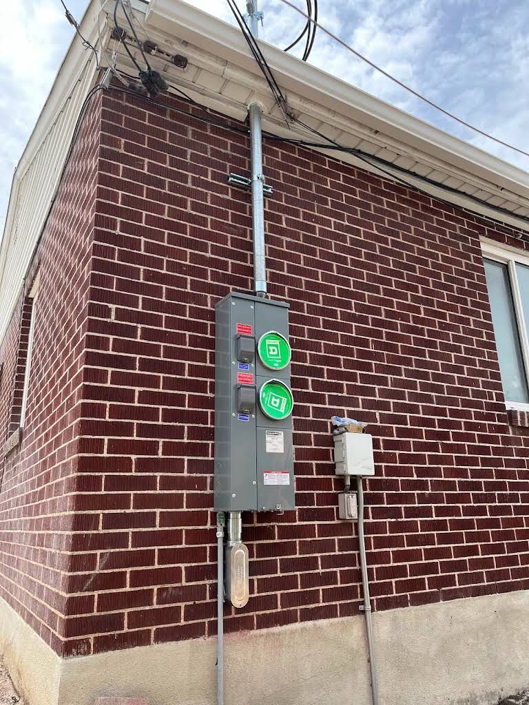 Exterior view of a brick building with a gray electrical meter box attached. Wires run along the building's side and top.