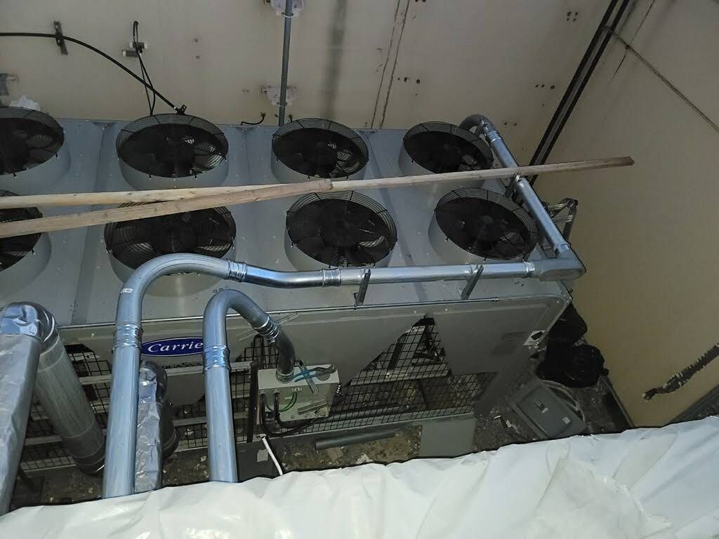 Large industrial cooling unit with six fan vents in a cramped, indoor setting. Gray metal, black fans, and white insulation are visible.