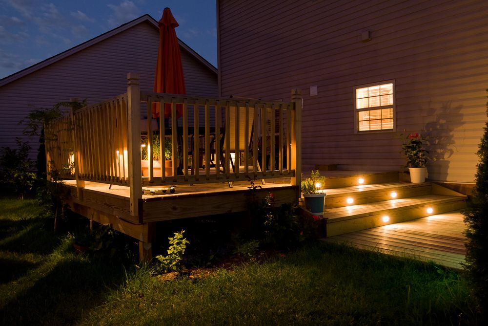 Lit-up wooden deck at dusk with stairs and house, warm lights, orange umbrella, and potted plants on the deck.