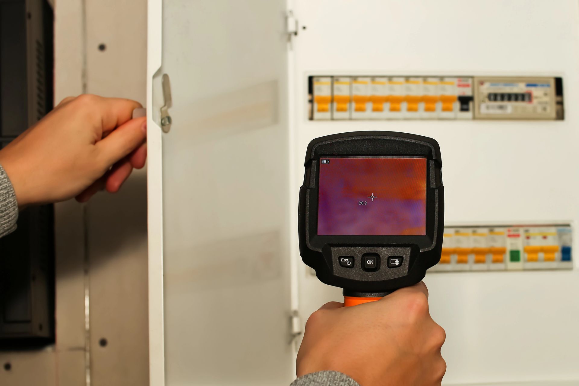 Person using a thermal camera on an electrical panel to check for overheating; indoor setting.