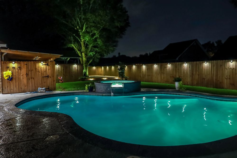 Nighttime backyard with illuminated pool, hot tub, wooden fence, and tree.
