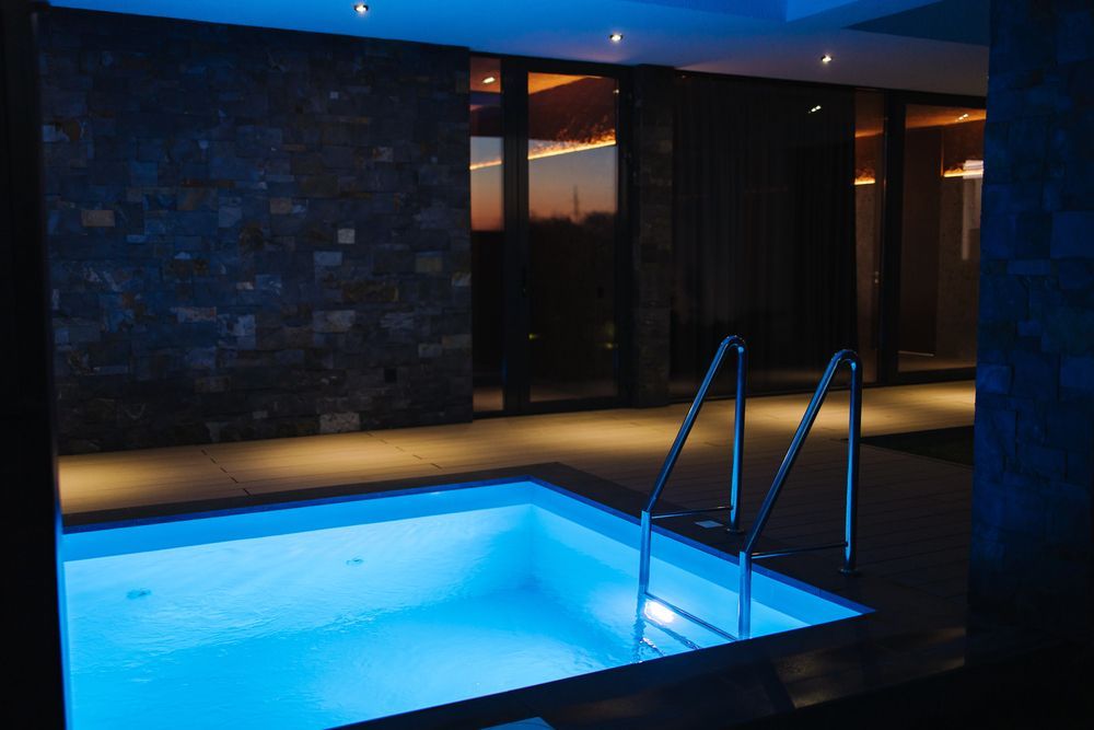 Indoor pool illuminated with blue light. Metal ladder, stone wall, and a sunset view through windows.