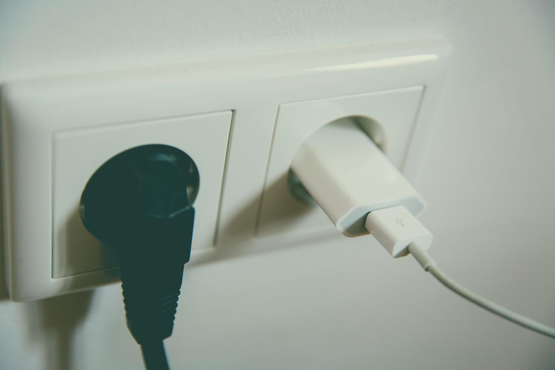 Two electrical outlets in a white wall. One has a black plug inserted, the other a white phone charger.