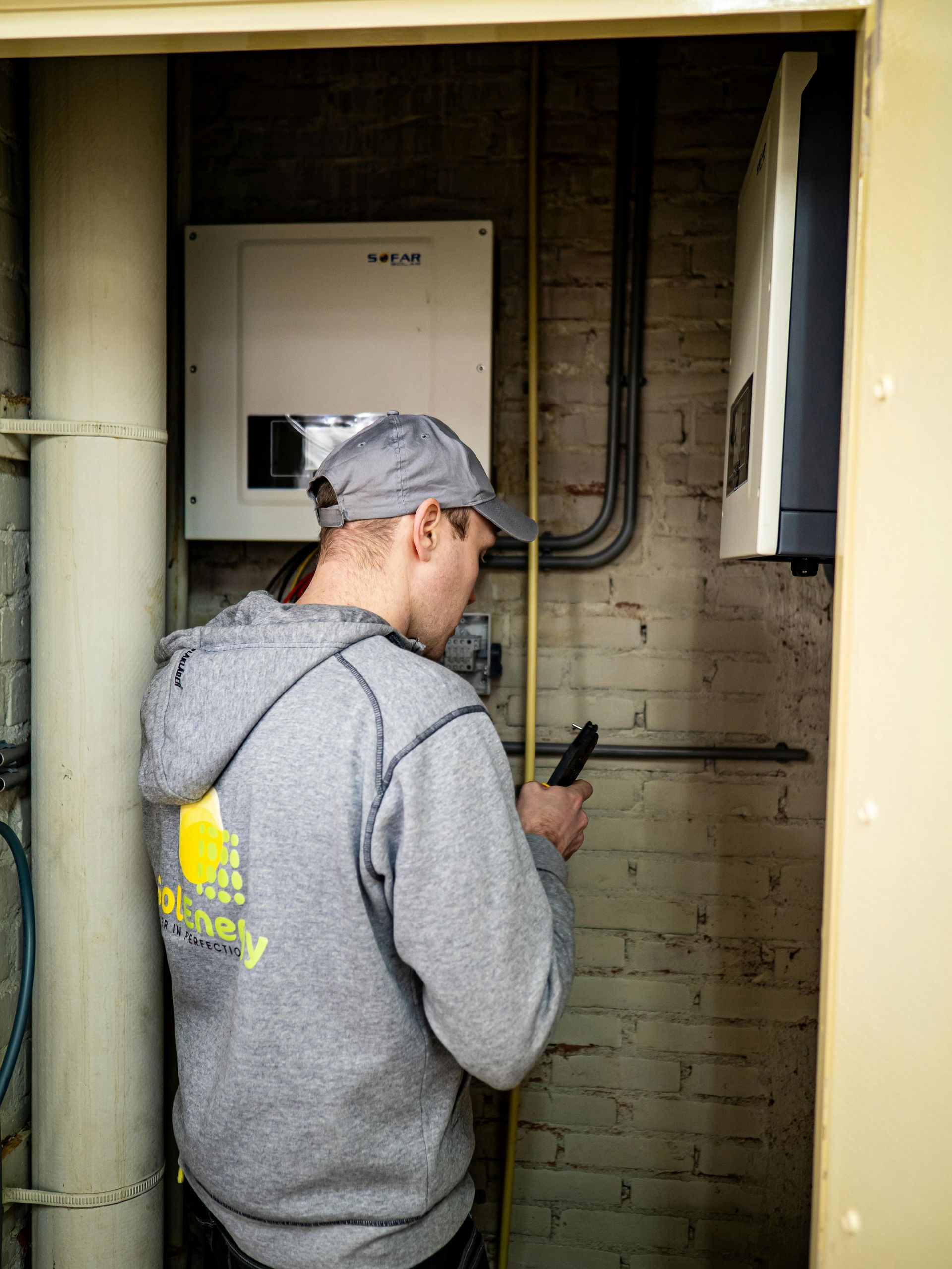 Man in gray hoodie and cap inspects electrical equipment in a utility closet. He holds a flashlight near the equipment and pipes.