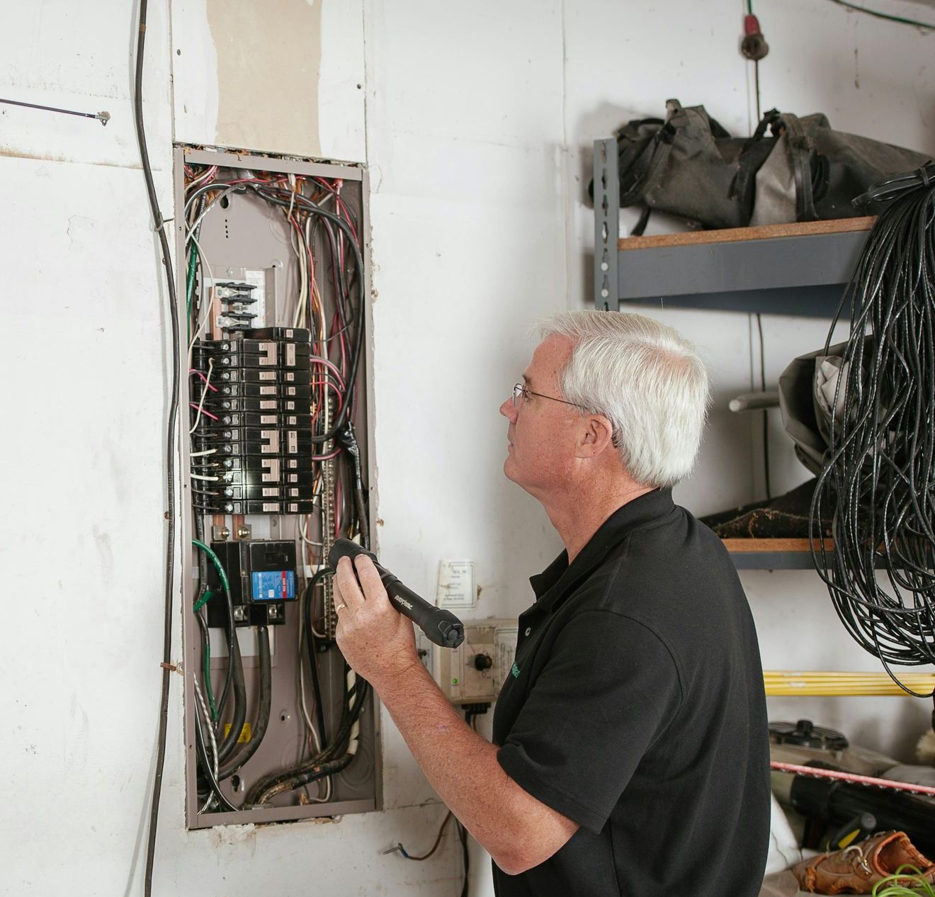 Electrician examines a circuit breaker panel with a flashlight in a garage.